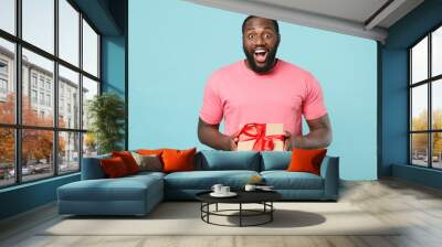 Surprised young african american man guy in pink t-shirt isolated on blue background. St. Valentine's Day, International Women's Day, birthday, holiday concept. Hold present box with gift ribbon bow. Wall mural