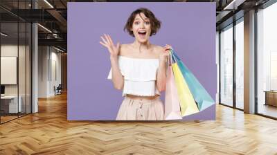 Surprised young woman girl in summer clothes hold package bag with purchases isolated on violet background. Shopping discount sale concept. Mock up copy space. Keeping mouth open, spreading hands. Wall mural