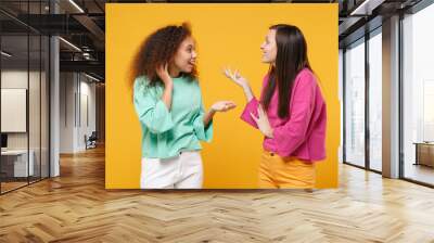 Two amazed women friends european and african american girls in pink green clothes posing isolated on yellow background. People lifestyle concept. Mock up copy space. Looking at each other speaking. Wall mural