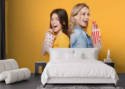 Two excited women girls friends in casual t-shirts denim clothes isolated on yellow background. People lifestyle concept. Mock up copy space. Hold cups of soda or cola, stand back to back, blinking. Wall mural