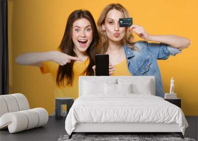 Two excited women girls friends in casual t-shirts denim clothes isolated on yellow background. People lifestyle concept. Pointing finger on mobile phone with blank empty screen hold credit bank card. Wall mural