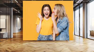 Two excited young women girls friends in casual t-shirts denim clothes isolated on yellow background. People lifestyle concept. Mock up copy space. Whispers gossip and tells secret with hand gesture. Wall mural