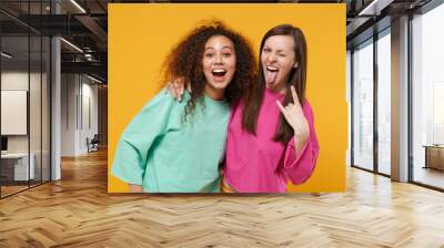 Two funny women friends european and african american girls in pink green clothes posing isolated on yellow orange background. People lifestyle concept. Mock up copy space. Hugging, showing horns up. Wall mural