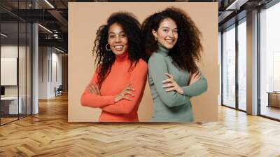 Two vivid smiling vivid young curly black women friends 20s wear casual shirts clothes stand back to back look at camera hold hands crossed isolated on plain pastel beige background studio portrait Wall mural