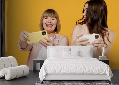 Two young excited daughter mother together couple women in casual clothes use play racing app on mobile cell phone hold gadget smartphone for pc video games isolated on plain yellow background studio Wall mural