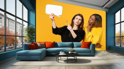 Two young women friends european and african american in black yellow clothes hold say clouds isolated on bright orange wall background, studio portrait. People lifestyle concept. Mock up copy space. Wall mural