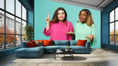 Two young women friends european and african american in pink green clothes standing posing isolated on blue turquoise wall background, studio portrait. People lifestyle concept. Mock up copy space. Wall mural