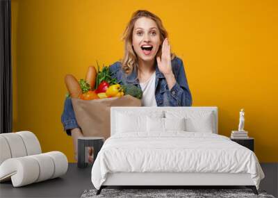 Woman in denim clothes isolated on orange background. Delivery service from shop or restaurant concept. Hold brown craft paper bag for takeaway mock up with food products screaming with hand gesture. Wall mural