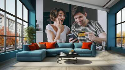 Young amazed couple two woman man in casual clothes sit by table eat pancake use mobile cell phone credit bank card shop online order delivery in light kitchen at home together Healthy diet concept Wall mural