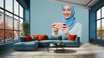 Young arabian asian muslim woman in abaya hijab using play racing app on mobile cell phone hold gadget smartphone isolated on plain blue background. People uae middle eastern islam religious concept. Wall mural