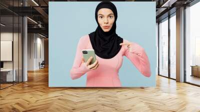 Young asian muslim fitness trainer sporty woman wear pink abaya hijab spend time in home gym use point finger mobile cell phone isolated on plain blue background studio. Workout sport fit abs concept. Wall mural