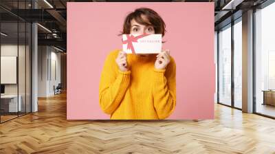 Young brunette woman in yellow sweater posing isolated on pastel pink background studio portrait. People sincere emotions lifestyle concept. Mock up copy space. Covering face with gift certificate. Wall mural