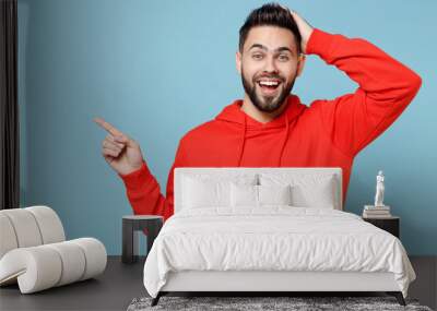 Young caucasian bearded surprised man in casual red orange hoodie hold head point index finger aside on copy space area mock up isolated on blue background studio portrait People lifestyle concept Wall mural