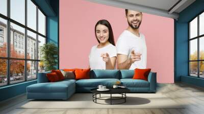 Young cheerful couple two friends bearded man brunette woman in white t-shirt looking camera standing back to back pointing finger camera isolated on pastel pink color background studio portrait Wall mural