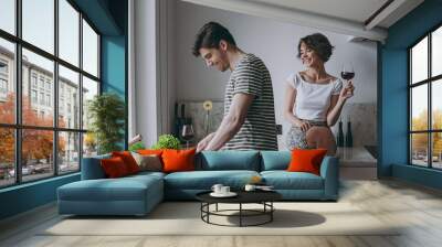 Young cheerful fun couple two woman man 20s in casual t-shirt clothes prepare vegetable salad girl sit table drink red wine cook food in light kitchen at home together. Healthy diet lifestyle concept Wall mural