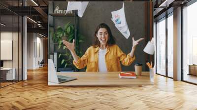 Young cheerful fun happy successful employee business woman 20s she wear casual yellow shirt throw up paper account documents sit work at wooden office desk with pc laptop. Achievement career concept. Wall mural