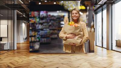 Young cheerful happy customer woman wear casual clothes look camera hold paper bag shopping at supermaket store grocery shop buying choose products in hypermarket. Purchasing food gastronomy concept. Wall mural