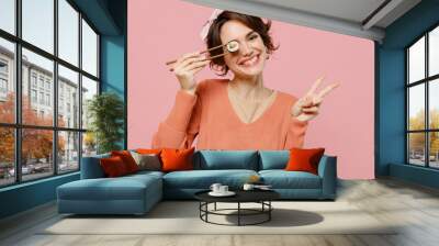 Young cheerful smiling happy fun cool woman in casual clothes hold in hand cover eye with makizushi sushi roll traditional japanese food showing victory sign isolated on plain pastel pink background Wall mural