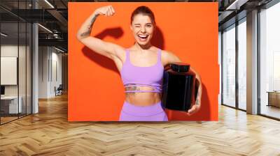 Young cool fitness trainer instructor woman sportsman wear purple top clothes in home gym hold bottle of supplements wink show biceps isolated on plain orange background Workout sport fit abs concept. Wall mural