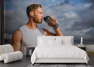 Young cool strong athletic toned fit sportsman man wears sports clothes earphones drink protein shake warm up train at sunrise sun dawn over sea beach outdoor on pier seaside in summer day morning.. Wall mural