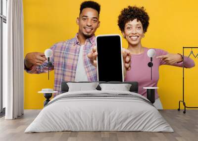 Young couple two friends family man woman of African American ethnicity wear purple casual clothes together use point finger on blank screen area mobile cell phone isolated on plain yellow background. Wall mural