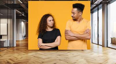 Young couple two friends together family offended frowning serious resentful african man woman in black tshirt look to each other hold hand crossed folded isolated on yellow background studio portrait Wall mural