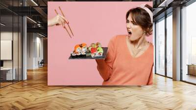 Young disturbed woman in sweater hold in hand makizushi sushi roll served on black plate traditional japanese food do not want share give to another person isolated on plain pastel pink background Wall mural