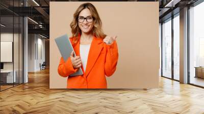 Young employee IT business woman corporate lawyer 30s in classic formal orange suit glasses work in office hold closed laptop pc computer point finger aside isolated on plain beige background studio Wall mural