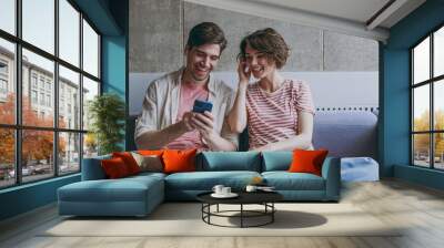 Young excited couple two friends woman man in casual clothes earphones sitting on sofa use mobile cell phone listen music radio online resting indoors at home flat together People lifestyle concept Wall mural