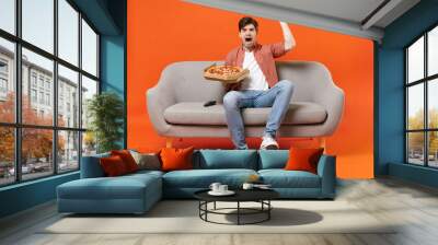 Young excited man football fan wear shirt support team with soccer ball sit on sofa at home watch tv live stream eat pizza do winner gesture isolated on orange background People sport leisure concept. Wall mural