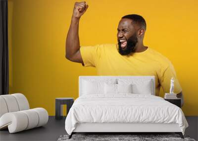 Young exultant happy black man 20s in bright casual t-shirt doing winner gesture celebrate clenching fists say yes isolated on plain yellow color background studio portrait. People lifestyle concept. Wall mural