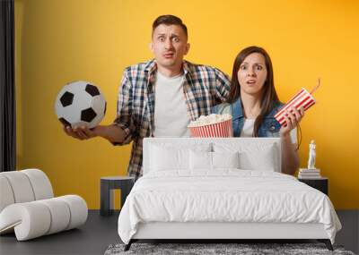 Young fun couple, woman man, football fans holding soccer ball, bucket of popcorn, plastic glass of cola, cheer up support team, isolated on yellow background. Sport family leisure lifestyle concept. Wall mural