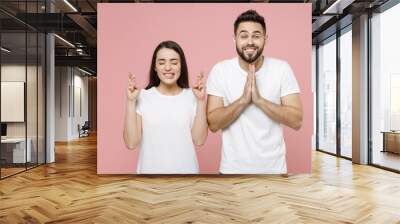 Young fun couple two friends man woman in white t-shirts wait for special moment keep fingers crossed making wish eyes closed hands folded in prayer gesture begging isolated on pastel pink background. Wall mural