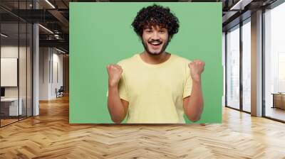 Young fun happy Indian man 20s in basic yellow t-shirt doing winner gesture celebrate clenching fists say yes isolated on plain pastel light green background studio portrait. People lifestyle concept. Wall mural