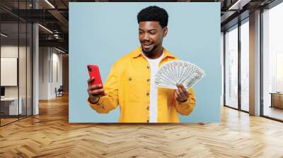 Young fun man of African American ethnicity 20s wear yellow shirt hold mobile cell phone fan of cash money in dollar banknotes isolated on plain pastel light blue background. People lifestyle concept. Wall mural