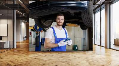 Young fun professional technician mechanic man wears denim blue overalls t-shirt use hold mobile cell phone stand near car lift check technical condition work in vehicle repair shop workshop indoors. Wall mural