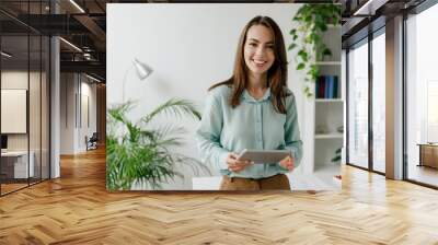 Young fun smiling european successful employee business woman in blue shirt work use hold tablet pc computer stand at workplace white desk at light modern office indoors. Achievement career concept. Wall mural