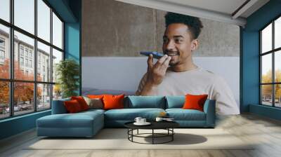 Young fun smiling happy african american man 20s wearing beige t-shirt sitting on grey sofa indoors apartment record voice message using mobile cell phone chatting resting on weekends staying at home. Wall mural