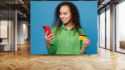 Young fun woman of African American ethnicity 20s wear green shirt using mobile cell phone hold credit bank card doing online shopping order delivery booking tour isolated on plain blue background. Wall mural