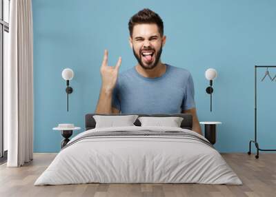 Young funny man in casual clothes posing isolated on blue background, studio portrait. People lifestyle concept. Mock up copy space. Showing tongue, depicting heavy metal rock sign, horns up gesture. Wall mural