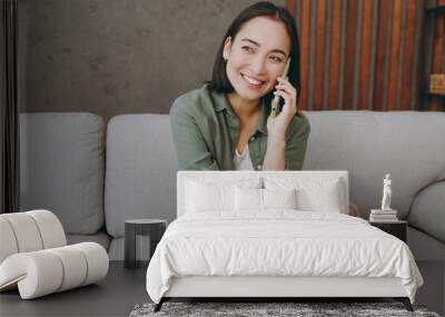 Young happy cheerful woman of Asian ethnicity wear casual clothes talk speak on mobile cell phone sit on grey sofa couch stay at home hotel flat rest relax spend free spare time in living room indoor Wall mural