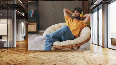 Young happy cool Indian man he wear orange casual clothes listen to music in headphones sits in armchair stay at home hotel flat rest relax spend free spare time in living room indoor. Lounge concept. Wall mural