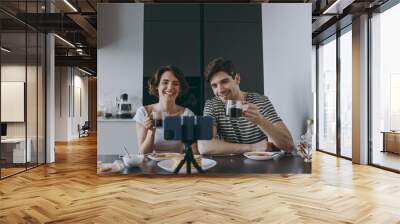 Young happy couple two woman man in casual t-shirt clothes sit by table eat pancakes talk video call mobile cell phone drinking coffee cook food in light kitchen at home together Healthy diet concept Wall mural