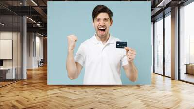 Young happy fun caucasian man 20s in white casual basic t-shirt hold in hand credit bank card do winner gesture clench fist isolated on pastel blue background studio portrait People lifestyle concept Wall mural