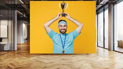 Young happy fun fitness trainer instructor sporty man sportsman in headband blue t-shirt golded medal hold winer cup above head isolated on plain yellow background. Workout sport motivation concept. Wall mural