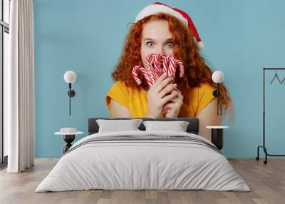 Young happy fun redhead woman 20s in yellow t-shirt santa claus red hat hold cover mouth with candy cane sweets isolated on plain light pastel blue background studio portrait People lifestyle concept Wall mural