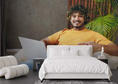 Young happy Indian IT man wear orange casual clothes hold use work point on laptop pc computer sits in armchair stay at home hotel flat rest relax spend free spare time in living room. Lounge concept. Wall mural