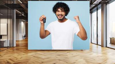 Young happy Indian man he wear white t-shirt casual clothes hold in hand car key fob keyless system do winner gesture isolated on plain pastel blue cyan background studio portrait. Lifestyle concept. Wall mural