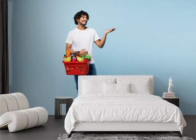 Young happy Indian man wear white t-shirt casual clothes hold red basket for takeaway mock up with food products point aside isolated on plain blue background Delivery service from shop or restaurant. Wall mural