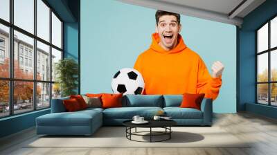 Young happy man fan he wears orange hoody casual clothes do winner gesture cheer up support football sport team hold in hand soccer ball watch tv live stream isolated on plain blue color background. Wall mural